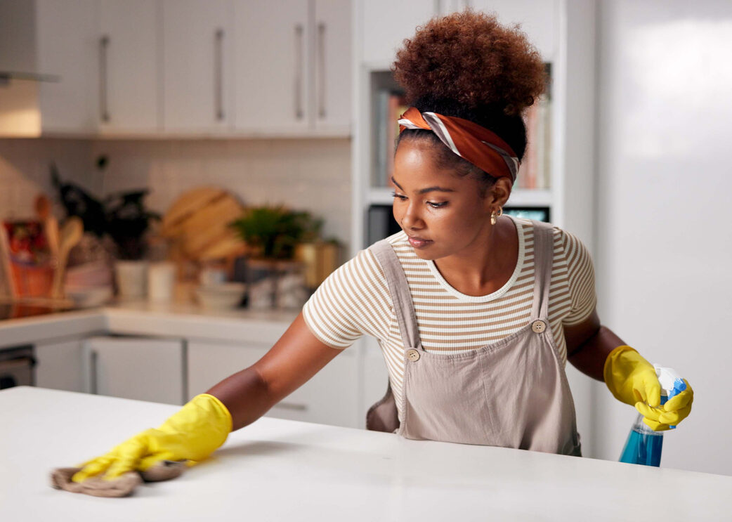 Woman, cloth and wipe on kitchen counter for hygiene with spray, gloves or chemical for shine in home. African person, cleaning and bottle for dust, dirt or bacteria with sanitation liquid in house